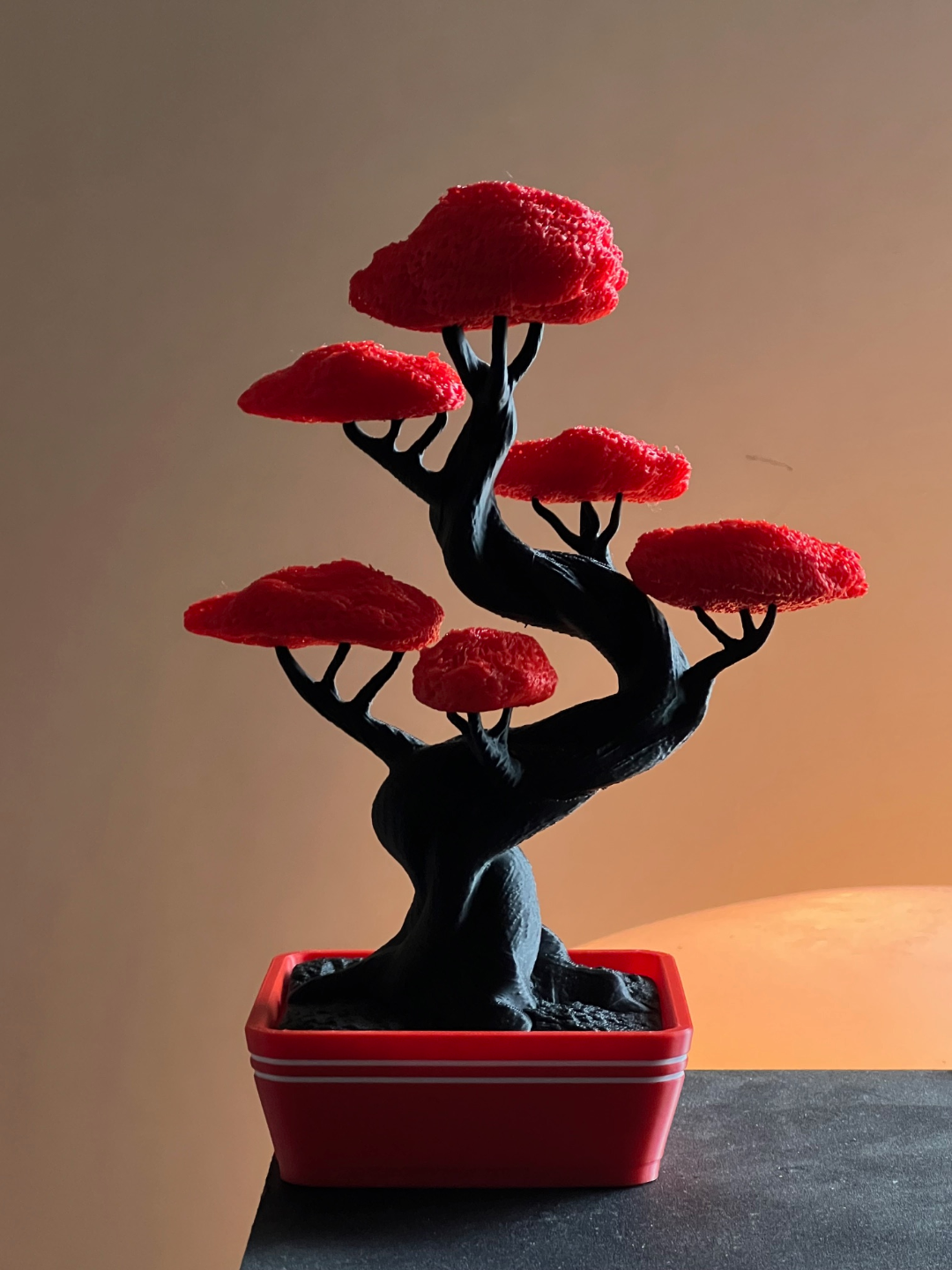 Figura impresa en 3D con forma de árbol bonsai en negro y rojo dentro de una maceta roja sobre un fondo cálido